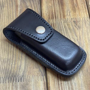 Könnte beinhalten: Eine schwarze Lederscheide mit silbernem Druckknopfverschluss. Die rechteckige Scheide mit abgerundeten Kanten weist detaillierte Nähte auf. Sie ist für ein kleines Werkzeug oder Messer konzipiert und etwa 15 cm lang.