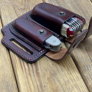 May include: A brown leather knife sheath with two pockets, each holding a multi-tool knife. The sheath has a belt loop and is secured with metal snaps. The knives have silver blades and red and silver handles.