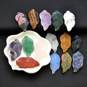 May include: Assortment of 14 leaf-shaped polished stones in various colors, including pink, green, purple, brown, white, orange, black, and blue. The stones are arranged on a white plate and a black background.