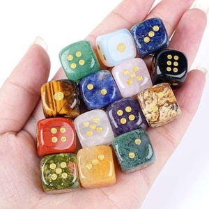 Peut inclure: Une collection de douze dés en pierres précieuses de différentes couleurs, dont vert, bleu, rouge et jaune. Chaque dé présente des points dorés. Les dés sont tenus dans une main, mettant en valeur leurs surfaces polies et leurs motifs uniques.