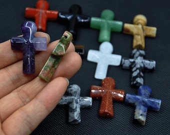 May include: A collection of gemstone crosses in diverse colours and textures. Includes red, black, green, tan, white, grey, orange, blue, and purple crosses. One cross is held in a hand, displaying the variety of materials and designs.