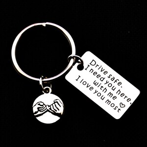 以下が含まれることがあります： 銀色のキーホルダーに、小指で約束をする2つの手の小さなチャームが付いています。チャームは、銀色のキーリングに付けられており、長方形のタグには「安全運転で。ここにいてほしい。私と一緒に。あなたを一番愛してる。」と書かれています。