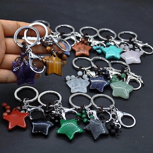 Puede incluir: Una colección de llaveros en forma de estrella en varios colores y materiales. Los llaveros presentan anillos y cierres plateados, con colgantes de estrella en tonos de rojo, negro, verde, morado, marrón y azul. Algunos tienen detalles de cuentas.