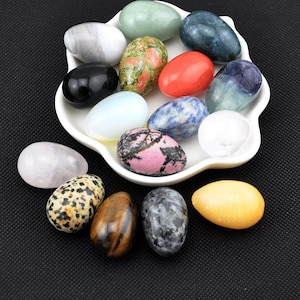 Op de afbeelding: Een verzameling van 15 gepolijste edelsteeneieren in verschillende kleuren en patronen, waaronder roze, groen, blauw, wit, zwart en bruin. De eieren worden gepresenteerd op een wit keramiek bord met een gegolfde rand.