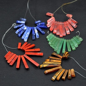 May include: A collection of gemstone beads in various colors, including blue, red, green, and brown. Each strand features rectangular-shaped beads strung together, creating a fan-like design. The beads are displayed against a dark background.