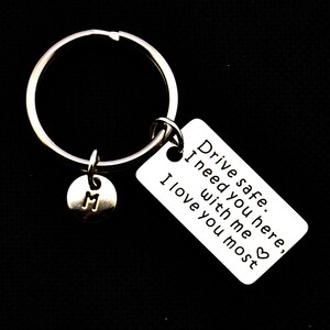 Op de afbeelding: Een zilverkleurige sleutelhanger met een grote ring, een kleine ronde bedel met de letter "M" en een rechthoekig label. Het label heeft de tekst "Drive safe. I need you here, with me, I love you most."