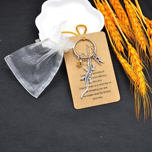 May include: A silver lizard charm keyring with an initial "R" charm and a small orange gem. The keyring is displayed on a brown card with text, next to a white organza bag and a decorative dish.