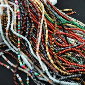 May include: A selection of gemstone beads in various colours and textures, including red, green, brown, black, and white. The beads are threaded onto thin cords and are arranged in a pile.