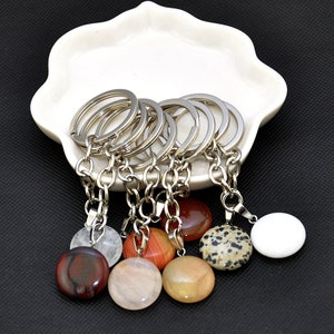 May include: A collection of keychains with round, polished gemstone pendants in various colors, including red, white, and brown. Each keychain features a silver-tone chain and keyring, displayed on a white, scalloped dish.