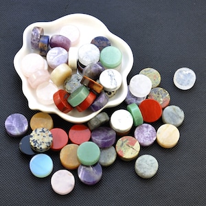 May include: Assortment of round, polished gemstone cabochons in various colours, including green, purple, pink, white, and brown. The stones are displayed in a white dish and scattered on a black surface.