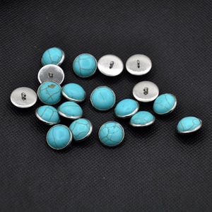 May include: A collection of turquoise and silver buttons. The buttons are round and have a raised, domed center. They are scattered on a black surface.
