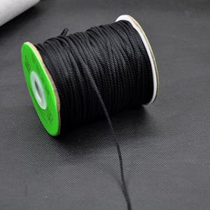 May include: A spool of black nylon cord. The cord is tightly wound around a cardboard core with green and white accents. The cord is unwound, with a single strand extending from the spool.