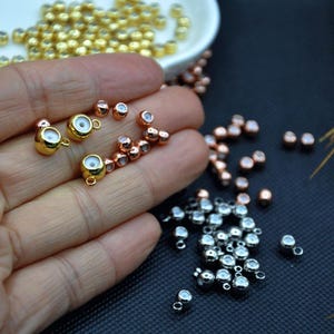 Op de afbeelding: Close-up van diverse sieraden-maken kralen in goud, koper en zilver tinten. De gouden kralen zijn kleine theepot-vormige bedels. Andere kralen zijn rond met een klein gaatje om te rijgen. Een hand houdt een aantal van de kralen vast.