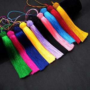 Puede incluir: Una colección de borlas coloridas en varios tonos, incluyendo verde, rosa, azul, amarillo y rojo. Cada borla presenta una parte superior anudada y una larga y fluida franja. Las borlas están dispuestas diagonalmente sobre un fondo oscuro.