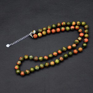 May include: A necklace featuring unakite gemstone beads in shades of green, orange, and red. The beads are strung on a green cord and secured with a silver-toned chain and clasp. The necklace is displayed against a dark background.