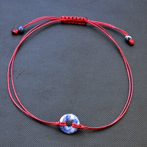 May include: A red string bracelet with a blue and white circle charm. The bracelet is tied with a knot at the top and has two small blue beads on either end.