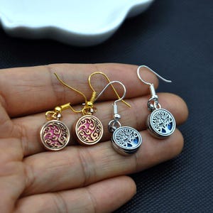 May include: Three pairs of earrings featuring a tree of life design. The earrings are silver, gold, and rose gold. The tree of life design is in the centre of each earring and is surrounded by a circle. The earrings hang from a hook.