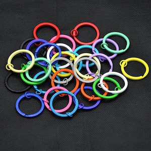 May include: Assortment of colourful metal key rings in various shades, including red, yellow, green, blue, and purple. The key rings are all approximately the same size and are arranged in a pile.