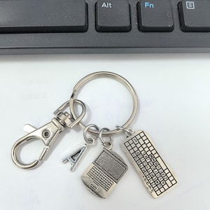 Puede incluir: Llavero plateado con un mosquetón, un dije con la letra "A" y dos dijes con temática de computadora: una computadora portátil y un teclado. El dije del teclado tiene letras y números.
