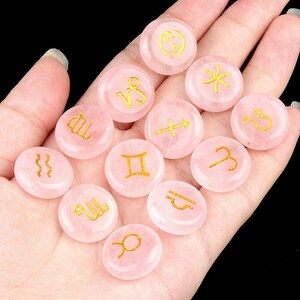 May include: Twelve pink rose quartz stones with gold zodiac symbols. The stones are smooth and round, and they are arranged in a circle.
