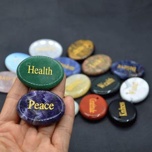 May include: Assortment of oval-shaped, smooth, polished stones with different colors and words inscribed in gold lettering. The words include "Health", "Peace", "Gratitude", "Wealth", "Serenity", "Wisdom", "Abundance", "Love", "Joy", "Freedom", "Strength", and "Harmony".