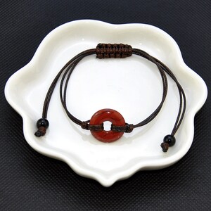 May include: A red stone bracelet with brown cord and black beads. The stone is round with a hole in the center.