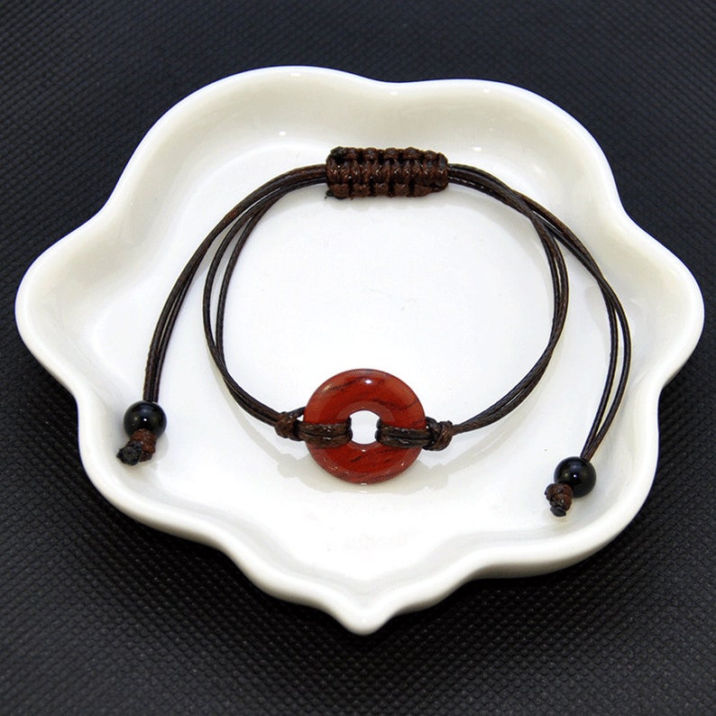 May include: A red stone bracelet with brown cord and black beads. The stone is round with a hole in the center.
