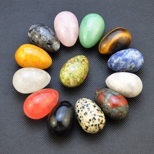 Peut inclure: Une collection de 12 œufs en pierres précieuses polies de différentes couleurs et motifs, notamment vert, bleu, rouge, noir, blanc et marron. Les œufs sont disposés en cercle sur une surface gris foncé.