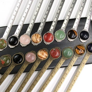 Könnte beinhalten: Eine Sammlung dekorativer Metalllineale mit Edelsteinakzenten. Jedes Lineal hat einen andersfarbigen Edelstein, darunter Schwarz, Rosa, Rot und Grün, der in einer dekorativen Metallfassung gefasst ist. Die Lineale werden auf einem schwarzen Tablett präsentiert.