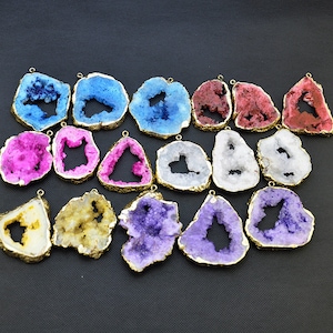 May include: A collection of geode pendants in various colors, including blue, pink, purple, and white. Each pendant has a gold-colored metal edge and a small loop for hanging. The geodes have a rough, textured surface and a hollow center.
