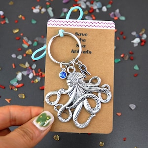 May include: Silver octopus keychain with a blue crystal charm and a silver letter 'R' charm. The keychain is attached to a silver key ring. The keychain is on a brown card with the text 'Save the Sea Animals'.