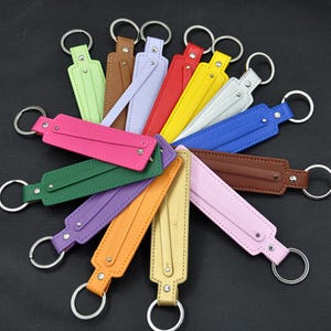 Op de afbeelding: Een verzameling kleurrijke leren sleutelhangers in verschillende tinten, waaronder groen, roze, paars en bruin. Elke sleutelhanger heeft een rechthoekige leren band met een metalen ring en klinknagels. De sleutelhangers zijn in een cirkelvorm gerangschikt.