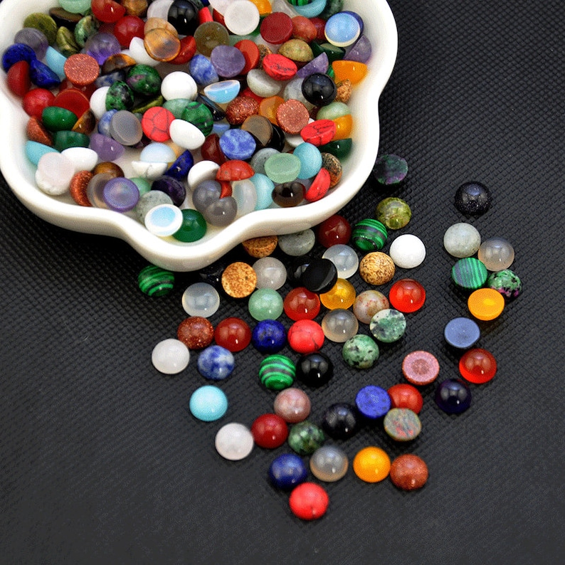 May include: A white bowl filled with a variety of colorful round gemstone cabochons. The bowl is on a black surface and some of the cabochons have spilled out.