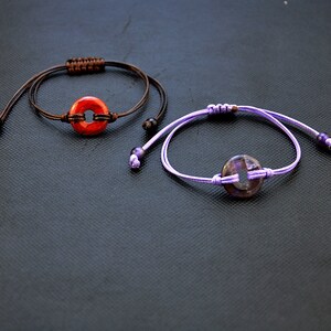 May include: Two bracelets with adjustable cords and round, flat, gemstone charms. The top bracelet has a brown cord and a red charm. The bottom bracelet has a purple cord and a purple charm.