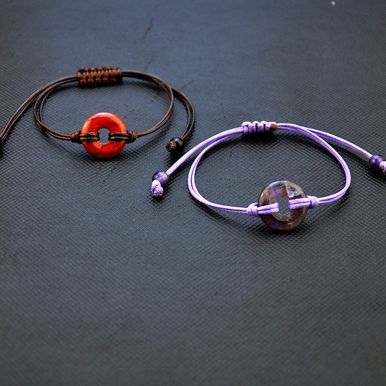May include: Two bracelets with adjustable cords and round, flat, gemstone charms. The top bracelet has a brown cord and a red charm. The bottom bracelet has a purple cord and a purple charm.
