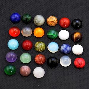 May include: Assortment of 25 round cabochon gemstones in various colors and patterns, including red, green, blue, white, and brown. The stones are arranged in a grid pattern on a black surface.