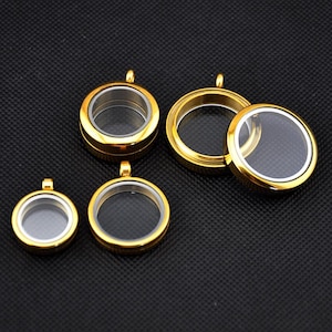 Könnte beinhalten: Mehrere goldfarbene, runde Anhänger-Medaillons mit transparenten Glaseinsätzen. Die Medaillons sind in verschiedenen Größen erhältlich, jedes mit einer kleinen Schlaufe für eine Kette. Ein Medaillon ist geöffnet. Der Hintergrund ist eine dunkle, strukturierte Oberfläche.