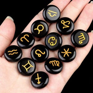 May include: Twelve black gemstone zodiac signs with gold lettering. The signs are arranged in a circle on a hand.