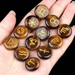May include: Twelve tiger's eye stones, each with a different zodiac symbol engraved in gold. The stones are arranged in a circle on a hand.