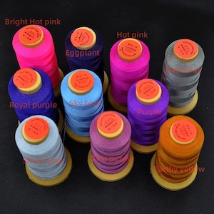 Puede incluir: Una colección de coloridas bobinas de hilo en tonos rosa, morado, azul, gris y amarillo. Cada bobina tiene una base de madera y una etiqueta redonda naranja. Los colores del hilo están etiquetados con sus nombres.