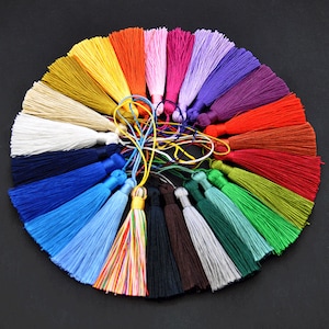 Puede incluir: Una colección de borlas coloridas dispuestas en un patrón circular. Las borlas están hechas de hilos sedosos en varios colores, incluyendo amarillo, naranja, rosa, morado y verde. Las borlas están atadas con cordones finos.