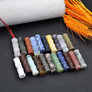 May include: A selection of cylindrical gemstone pendants, each with a bamboo-like design. The pendants are in various colours, including green, blue, brown, and white. A red string is attached to one pendant. The pendants are displayed on a black surface.