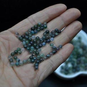 May include: A close-up of a hand holding a pile of green and white agate beads. The beads are round and have a smooth, polished finish.