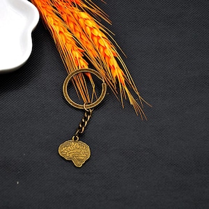 Può includere: Un portachiavi color bronzo con un ciondolo a forma di cervello. Il portachiavi ha un anello circolare e una catena corta che collega il ciondolo a forma di cervello. Lo sfondo include spighe di grano arancioni e una superficie scura.