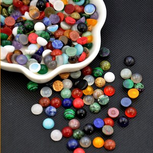 May include: A white bowl filled with a variety of colorful round gemstone cabochons. The bowl is on a black surface and some of the cabochons have spilled out of the bowl.
