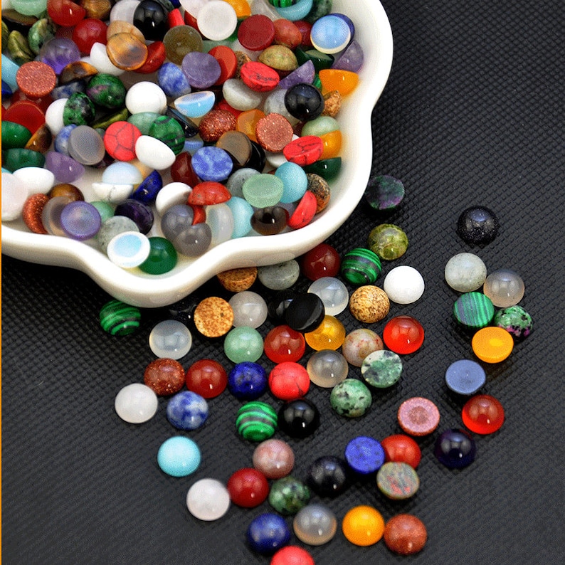May include: A white bowl filled with a variety of colorful round gemstone cabochons. The bowl is on a black surface and some of the cabochons have spilled out of the bowl.