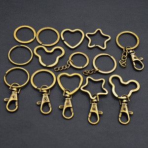 May include: A collection of antique brass-colored keychains in various shapes, including circles, hearts, stars, and a Mickey Mouse-like design. Some keychains have a lobster clasp and chain. The keychains are arranged on a dark background.