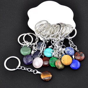 May include: A collection of keychains with round, polished gemstones in various colors, including green, purple, blue, and brown. Each keychain has a silver-colored ring and chain. The gemstones are displayed on a white, cloud-shaped dish.
