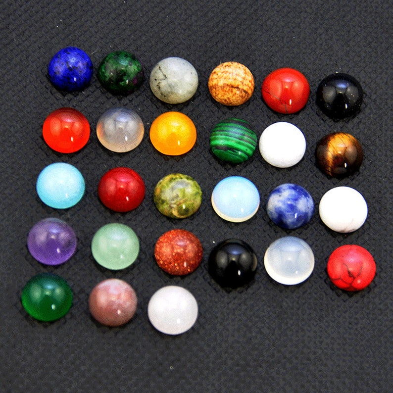 May include: Assortment of 25 round cabochon gemstones in various colors and patterns, including red, green, blue, white, and brown. The gemstones are arranged in a grid pattern on a black surface.
