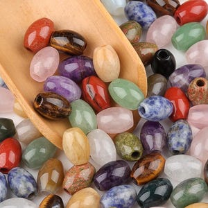 Peut inclure: Un assortiment de perles de pierres précieuses de forme ovale, de couleurs variées, dont le rouge, le vert, le violet et le bleu. Les perles sont présentées dans une cuillère en bois, mettant en valeur leurs surfaces polies et leurs variations naturelles. Ces perles sont adaptées à la création de bijoux.